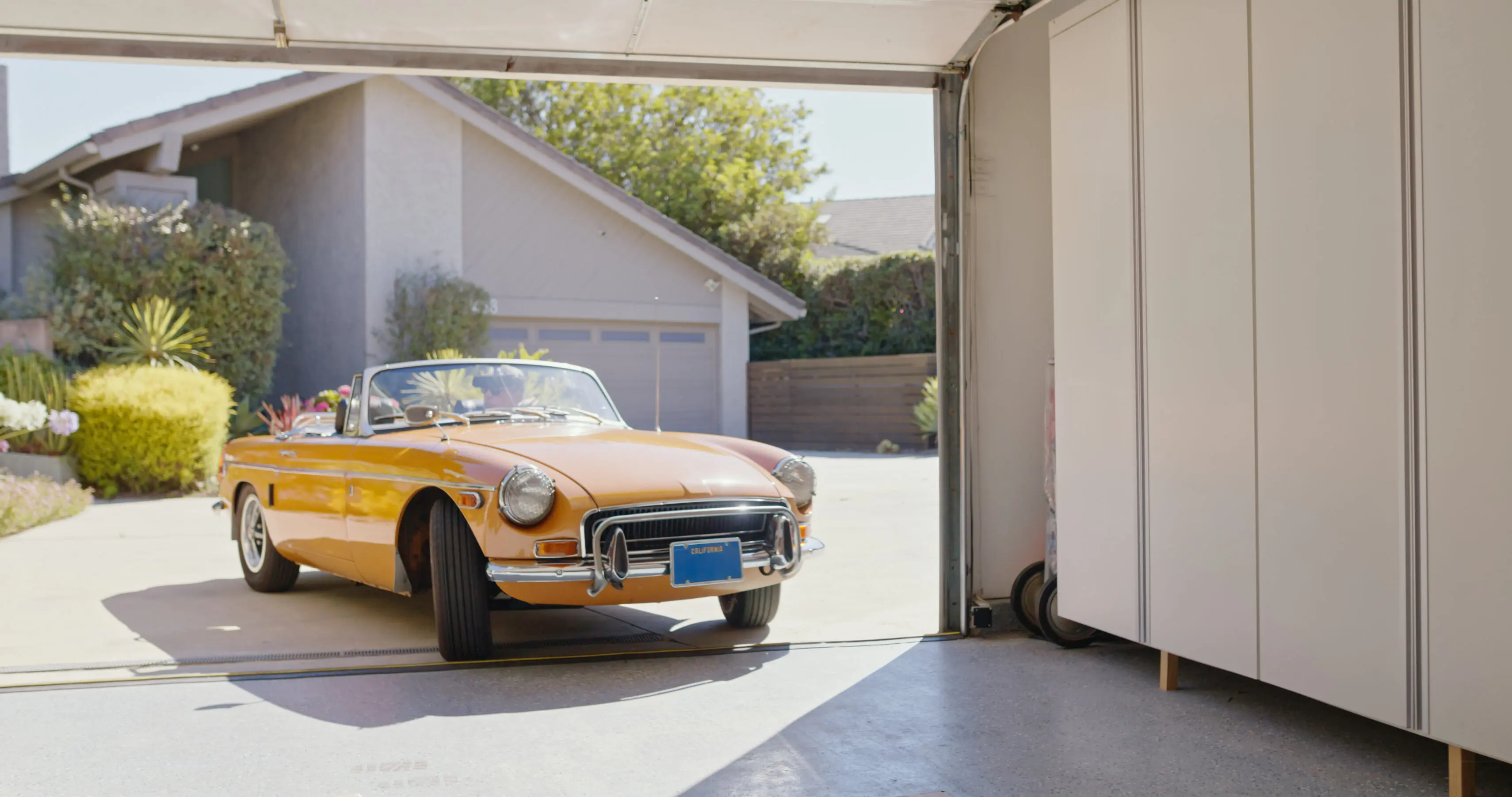 A wide shot of a vintage car backing out of a garage and driving away on a sunny day.